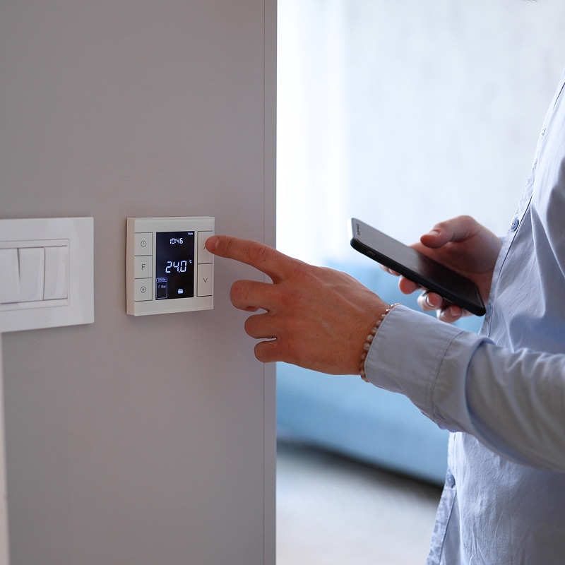 Thermostat intelligent : réglage de la température avec le mobile Homme réglant un thermostat intelligent mural affichant 24.0°C et 10:46, tenant un smartphone noir.